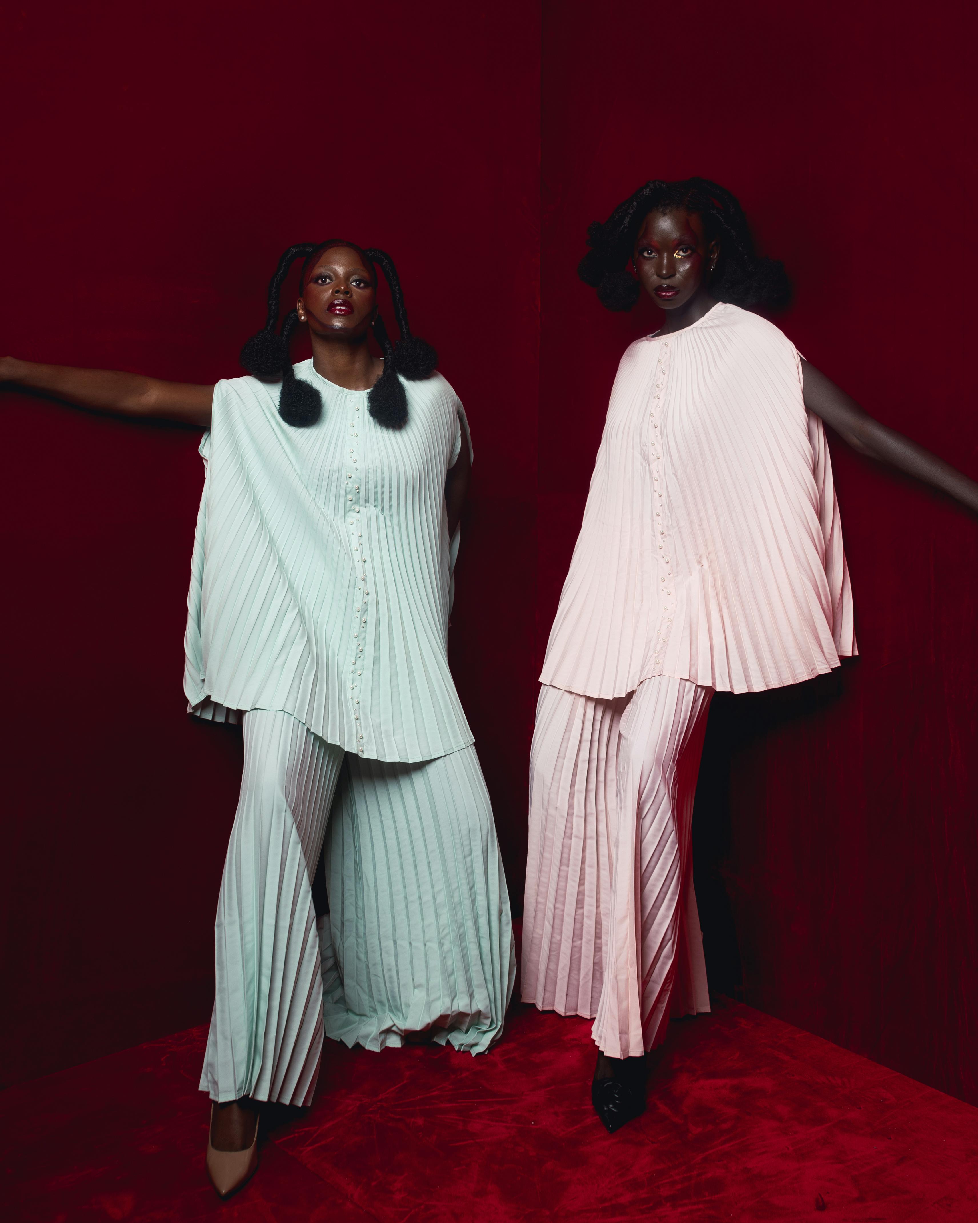 Two girls with unique hair styles in front of a deep red background and pleated outfits that are the same but different colors one light green and the other light pink