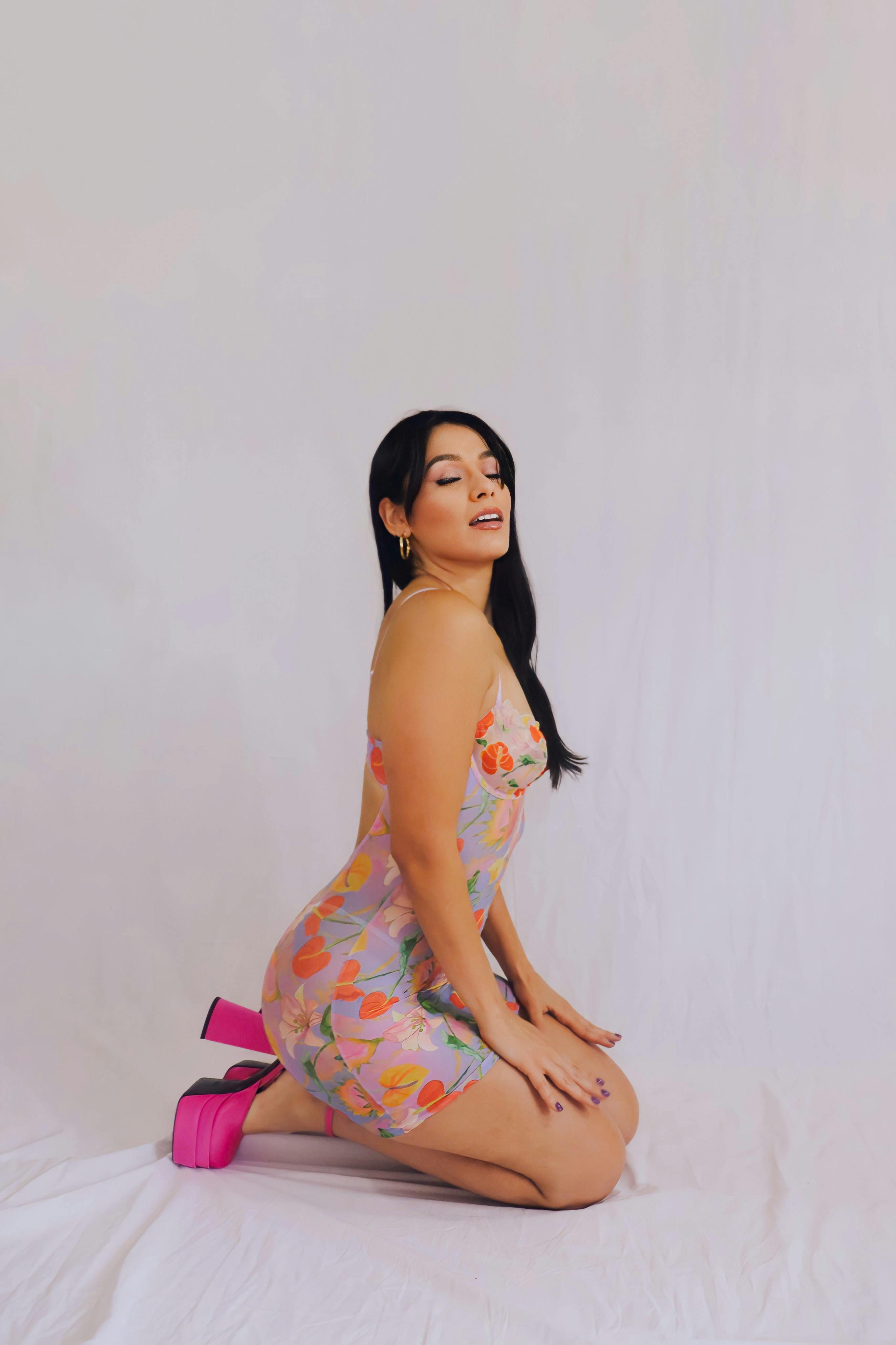 Editorial image of a girl in a floral dress with pink heels on her knees in front of a white sheet wall