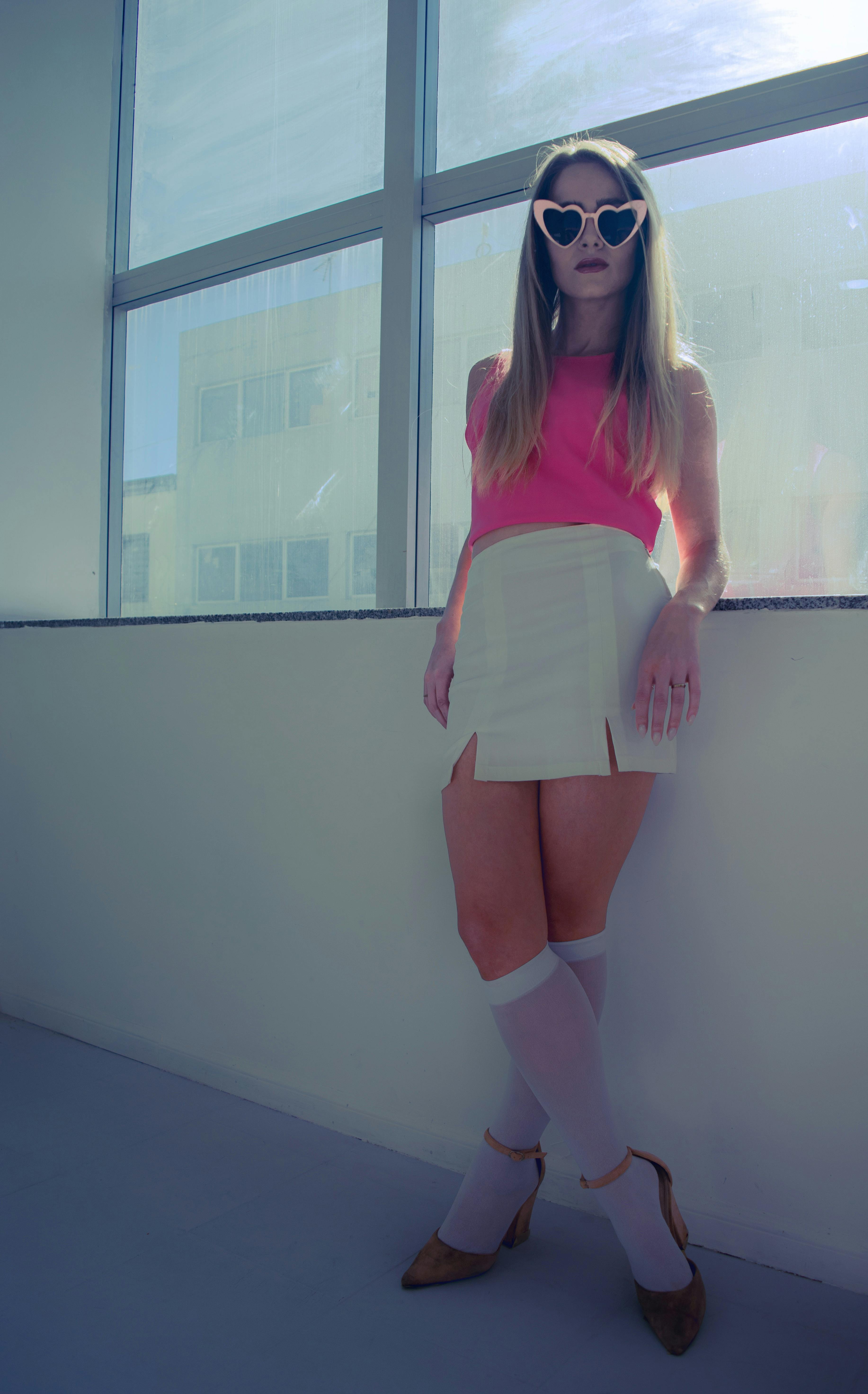 Girl with heart sunglasses standing in front a window leaning on the ledge in a white mini skirt and pink tank top